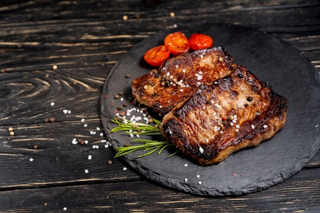 A juicy piece of fried meat with grilled cherry tomatoes lies on a stone plate against a black wooden table. Degree of roasting well done. Food concept with copy spaceの写真素材