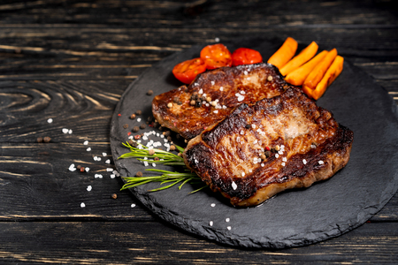 A juicy piece of fried meat with grilled cherry tomatoes and carrot lies on a stone plate against a black wooden table. Degree of roasting well done. Food concept with copy spaceの写真素材