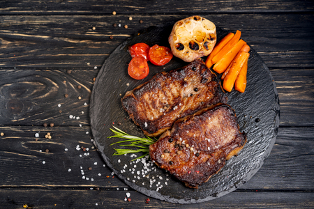 A juicy piece of fried meat with grilled cherry tomatoes garlic and carrot lies on a stone plate against a black wooden table. Degree of roasting well done. Food concept with copy spaceの写真素材