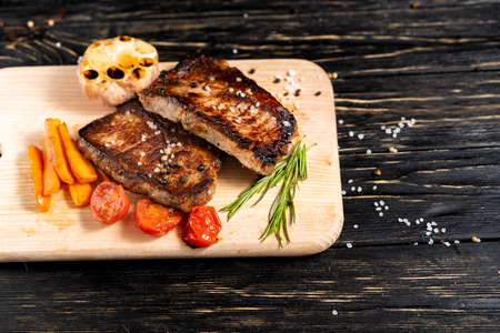 A juicy piece of fried meat with grilled cherry tomatoes garlic and carrot lies on a cutting board against a black wooden table. Degree of roasting well done. Food concept with copy spaceの写真素材