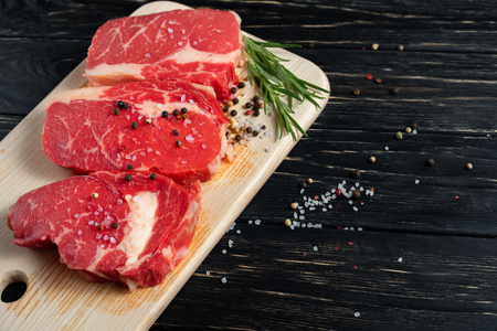 Three pieces of juicy raw beef with rosemary on a cutting board on a black wooden table background. Meat for barbecue or grill sprinkled with pepper and salt seasoningの写真素材