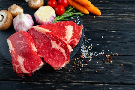 Three pieces of juicy raw beef with rosemary on a stone cutting board on a black wooden table background. Meat for barbecue or grill with vegetables pepper and salt seasoningの写真素材