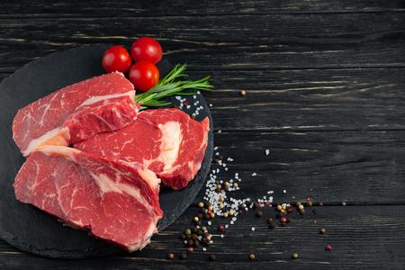 Three pieces of juicy raw beef with rosemary on a stone cutting board on a black wooden table background. Meat for barbecue or grill next to cherry tomatoes sprinkled with pepper and salt seasoningの写真素材