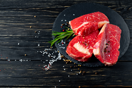 Three pieces of juicy raw beef with rosemary on a stone cutting board on a black wooden table background. Meat for barbecue or grill sprinkled with pepper and salt seasoningの写真素材