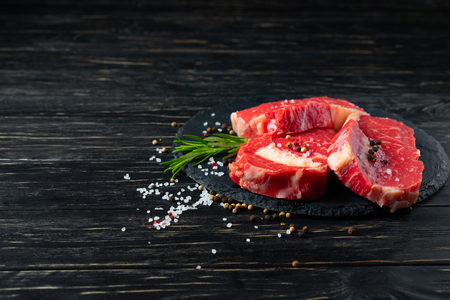 Three pieces of juicy raw beef with rosemary on a stone cutting board on a black wooden table background. Meat for barbecue or grill sprinkled with pepper and salt seasoningの写真素材