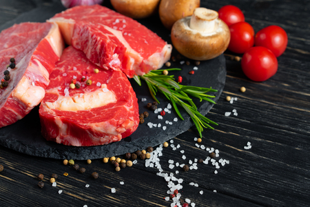 Three pieces of juicy raw beef with rosemary on a stone cutting board on a black wooden table background. Meat for barbecue or grill with tomatoes mushrooms pepper and salt seasoningの写真素材