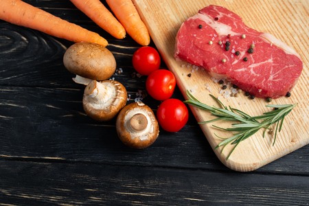One pieces of juicy raw beef with rosemary on a cutting board on a black wooden table background. Meat for barbecue or grill with vegetables pepper and salt seasoningの写真素材