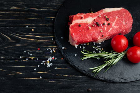 One pieces of juicy raw beef with rosemary on a stone cutting board on a black wooden table background. Meat for barbecue or grill next to cherry tomatoes sprinkled with pepper and salt seasoningの写真素材