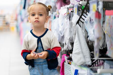 A little Caucasian girl is standing near a shop window with clothes and underwear in a large store. Little buyer. Consumption generationの写真素材