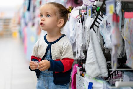 A little Caucasian girl is standing near a shop window with clothes and underwear in a large store. Little buyer. Consumption generationの写真素材