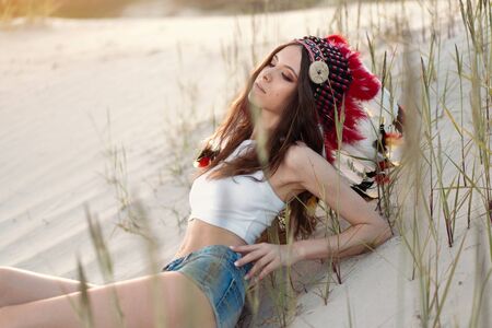 A beautiful young Caucasian girl in a white top on her head is wearing an Indian hat Roach Lies in the desert. close up portraitの写真素材