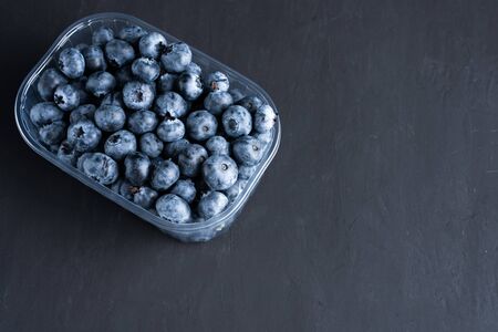 Tasty juicy raw blueberries in a plastic container on a black dark background. Packaging for berries...
