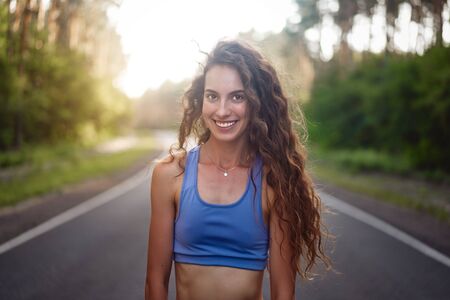 close up portrait Beautiful young Caucasian girl standing on asphalt forest road smiling before jogging. Athlete before jogging. Morning workout nature. Sport motivation and healthy lifestyle conceptの写真素材