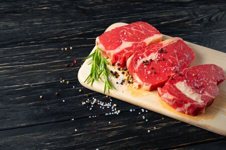 Three pieces of juicy raw beef with rosemary on a cutting board on a black wooden table background. Meat for barbecue or grill sprinkled with pepper and salt seasoningの写真素材