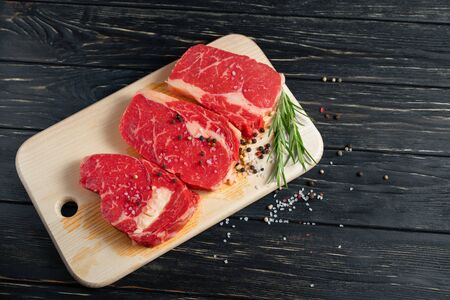 Three pieces of juicy raw beef with rosemary on a cutting board on a black wooden table background. Meat for barbecue or grill sprinkled with pepper and salt seasoningの写真素材