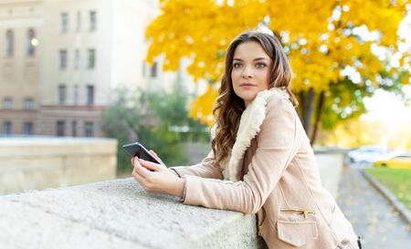 Beautiful caucasian brunette girl standing warm autumn day with background of trees with yellow foliage and a city. Dressed wool sweater and jacket. Holds mobile phone in handの写真素材