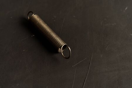 A metal spring with fasteners at the ends lies on a black cement background.