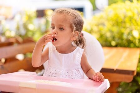 Cute little Caucasian girl eating spaghetti at table sitting in child seat outdoor restaurant. Healthy eating. Funny child eat street food sunny summer day.の写真素材