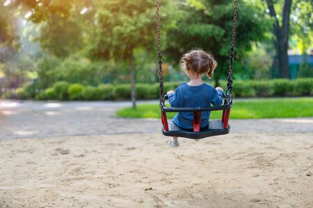 Unrecognizable Little Caucasian girl riding swing at playground sunny summer day rear view. European child having fun outdoor. Family leisure on park. Funny child enjoy the summertime.の写真素材