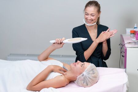 Cosmetology cabinet client lies on couch looks mirror result of the work done Beautician smiles looking at the clients. Positive emotion. Cosmetologist do well job. Free space. Beauty industryの写真素材