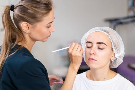 Cosmetology cabinet client sitting on couch. Beautician applies marking on eyebrows. Preparation for the procedure of permanent eyebrow makeup. Free space. Beauty industryの写真素材