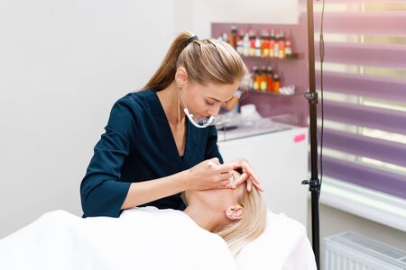 Cosmetology cabinet client lies on couch. Beautician applies marking on eyebrows. Preparation for the procedure of permanent eyebrow makeup. Free space. Beauty industryの写真素材