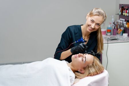 Cosmetology cabinet client lies on couch. Beautician applies permanent ink on eyebrows. Procedure of permanent eyebrow makeup. Free space. Beauty industryの写真素材