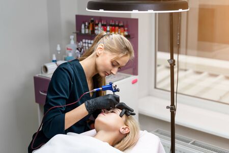 Cosmetology cabinet client lies on couch. Beautician applies permanent ink on eyebrows. Procedure of permanent eyebrow makeup. Free space. Beauty industryの写真素材