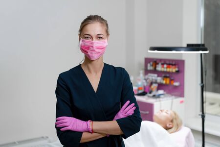 Cosmetology cabinet client lies on couch. Beautician stands in pink medical mask and smiles. Preparation for the procedure of permanent eyebrow makeup.の写真素材
