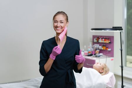 Cosmetology cabinet client lies on couch. Beautician stands in pink medical mask and smiles. Preparation for the procedure of permanent eyebrow makeup.の写真素材