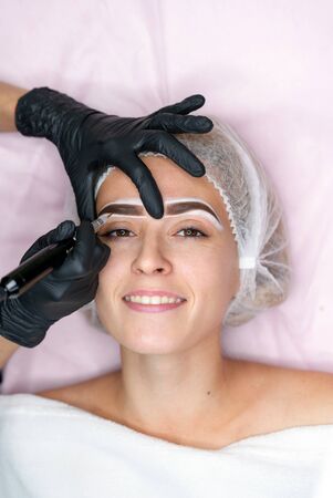 Cosmetology cabinet client lies on couch. Beautician applies permanent ink on eyebrows. Procedure of permanent eyebrow makeup. Close up black glove. Beauty industryの写真素材