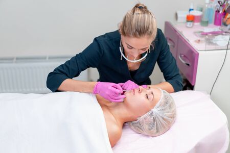 Cosmetology cabinet client lies on couch. Beautician applies permanent ink on lips. Procedure of permanent lip makeup. Free space. Beauty industryの写真素材