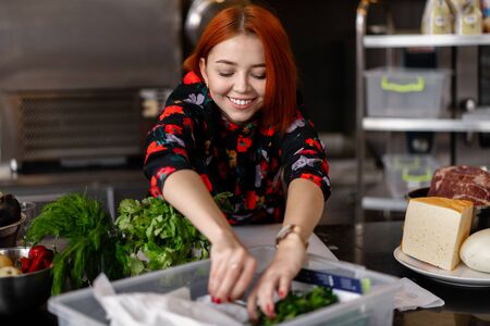 Beautiful young red-haired girl in evening dress is preparing food in a professional kitchen. Cheereful Woman making salad and cooking dinner for many peopleの写真素材