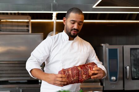 unrecognizable young African chef standing in professional kitchen in restaurant preparing a meal of meat and cheese vegetables. Portrait of man in cook uniform Cuts meatの写真素材