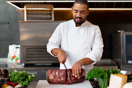 Handsome young African chef standing in professional kitchen in restaurant preparing a meal of meat and cheese vegetables. Portrait of man in cook uniform Cuts meat with a metal knife.の写真素材