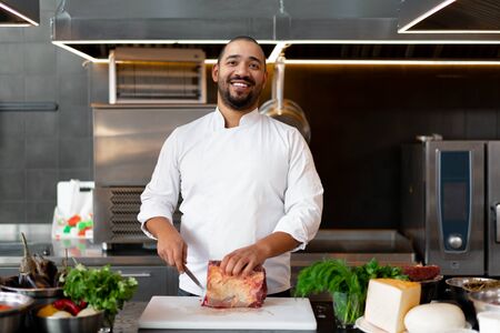Handsome young African chef standing in professional kitchen in restaurant preparing a meal of meat and cheese vegetables. Portrait of man in cook uniform Cuts meat with a metal knife.の写真素材