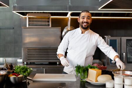 Handsome young African chef standing in professional kitchen in restaurant preparing a meal of meat and cheese vegetables. Portrait of man in cook uniform. Healthy eating concept.の写真素材