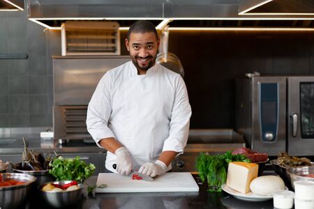 Handsome young African chef standing in professional kitchen in restaurant preparing a meal of meat and cheese vegetables. Portrait of man in cook uniform Cuts red pepper with a metal knife.の写真素材