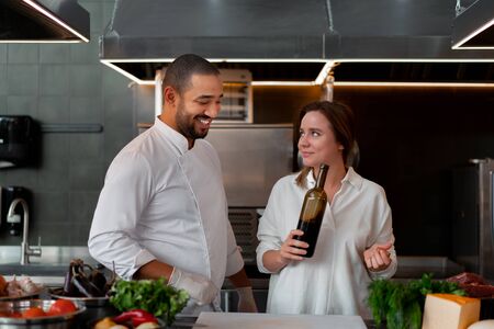 Handsome young African chef is cooking together with Caucasian girlfriend in the kitchen using red wine ingredient. A cook teaches a girl how to cook. Man and woman cooking in professional kitchen.の写真素材