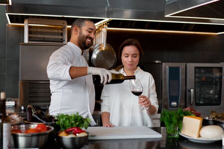 Handsome young African chef is cooking together with Caucasian girlfriend in the kitchen using red wine ingredient. A cook teaches a girl how to cook. Man and woman cooking in professional kitchen.の写真素材