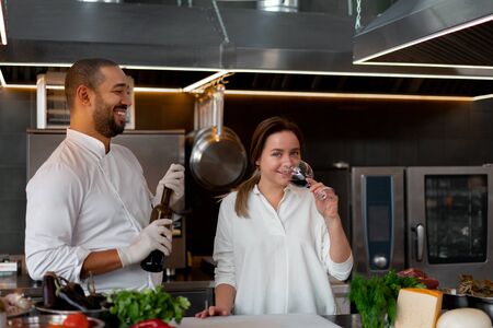Handsome young African chef is cooking together with Caucasian girlfriend in the kitchen using red wine ingredient. A cook teaches a girl how to cook. Man and woman cooking in professional kitchen.の写真素材