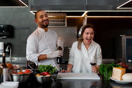 Handsome young African chef is cooking together with Caucasian girlfriend in the kitchen using red wine ingredient. A cook teaches a girl how to cook. Man and woman cooking in professional kitchen.の写真素材