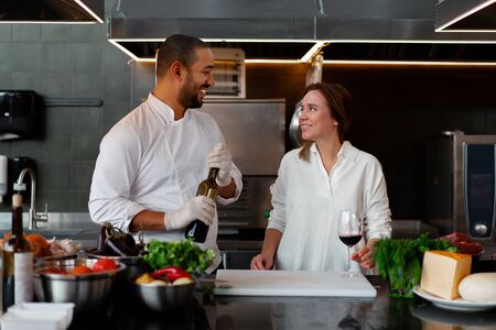 Handsome young African chef is cooking together with Caucasian girlfriend in the kitchen using red wine ingredient. A cook teaches a girl how to cook. Man and woman cooking in professional kitchen.の写真素材