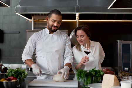 Handsome young African chef is cooking together with Caucasian girlfriend in the kitchen using red wine ingredient. A cook teaches a girl how to cook. Man and woman cooking in professional kitchen.の写真素材