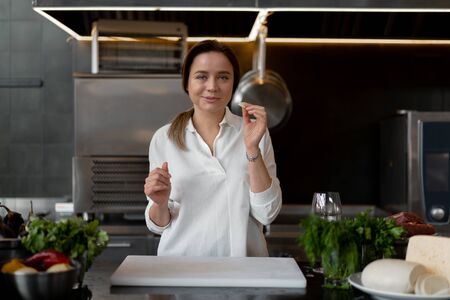 Beautiful young caucasian girl standing in kitchen in a white uniform smiling and tasting a piece of cheese. Cute woman 30s years old in white shirt with food Ingredients cheese meat vegetables.の写真素材
