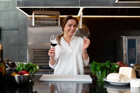 Beautiful young caucasian girl standing in kitchen in a white uniform smiling and tasting red wine Cute woman 30s years old in white shirt with food Ingredients cheese meat vegetables drinkingの写真素材