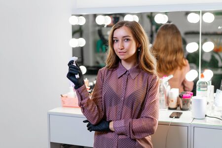 Beautiful young Caucasian makeup artist standing with tool in hands on background mirror in a beauty salon. Portrait of beautician at his workplace ready to work.の写真素材