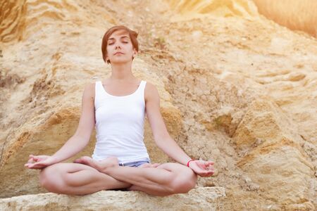 A beautiful young girl with short hair is dressed in shorts and a white jersey is practicing yoga on the background of rocks. Pose of the lotus. The concept of calm and concentration.の写真素材