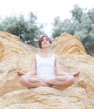 A beautiful young girl with short hair is dressed in shorts and a white jersey is practicing yoga on the background of rocks. Pose of the lotus. The concept of calm and concentration.の写真素材