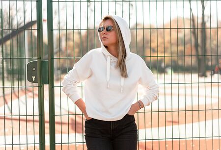 Outdoor close up portrait of young beautiful woman with long hair in sunglasses, dressed in a white hoodie sweater, near the sportsground. youth culture summer pastimeの写真素材